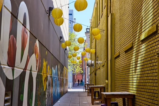 Vibrant urban alley in Fort Wayne with a colorful street mural, festive yellow lanterns, and cozy dining setup.