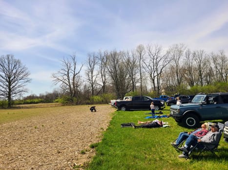 Spring outing as Eclipse darkens the skies in rural Indiana: Adults relax and child plays in a lush, green field under a clear sky.