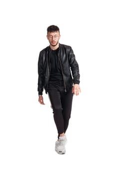 Full-length photo of a modern performer in glasses, black leather jacket, t-shirt, sports pants and light sneakers fooling around in studio. Indoor photo of a comical guy dancing isolated on white background. Music and imagination.