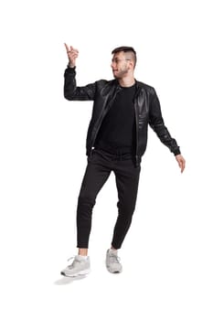 Full-length photo of a modern performer in glasses, black leather jacket, t-shirt, sports pants and light sneakers fooling around in studio. Indoor photo of a comical person dancing and looking away isolated on white background. Music and imagination.