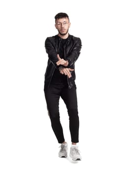Full-length photo of a modern performer in glasses, black leather jacket, t-shirt, sports pants and light sneakers fooling around in studio. Indoor photo of a stylish fellow dancing and gesticulating isolated on white background. Music and imagination.