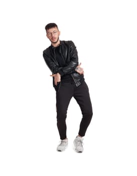 Full-length photo of a modern performer in glasses, black leather jacket, t-shirt, sports pants and light sneakers fooling around and gesticulating in studio. Indoor photo of an intelligent fellow dancing isolated on white background. Music and imagination.
