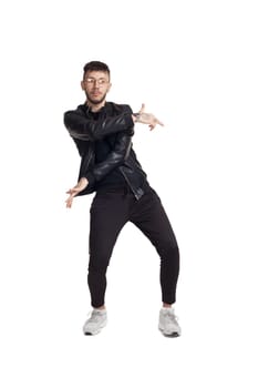 Full-length photo of a modern performer in glasses, black leather jacket, t-shirt, sports pants and light sneakers fooling around in studio. Indoor photo of a mischievous fellow dancing and gesticulating isolated on white background. Music and imagination.