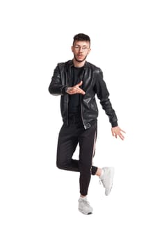 Full-length photo of a modern performer in glasses, black leather jacket, t-shirt, sports pants and light sneakers fooling around in studio. Indoor photo of a mischievous man dancing and looking at the camera isolated on white background. Music and imagination.