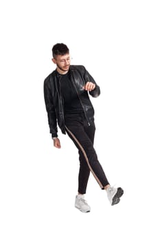 Full-length photo of a modern performer in glasses, black leather jacket, t-shirt, sports pants and light sneakers fooling around in studio. Indoor photo of a strong fellow dancing and looking down isolated on white background. Music and imagination.
