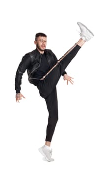 Full-length photo of a modern performer in glasses, black leather jacket, t-shirt, sports pants and light sneakers fooling around in studio. Indoor photo of a strong male dancing and raising his leg isolated on white background. Music and imagination.