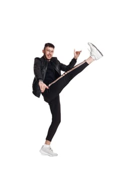 Full-length photo of a modern performer in glasses, black leather jacket, t-shirt, sports pants and light sneakers fooling around in studio. Indoor photo of a funny person dancing and raising his leg isolated on white background. Music and imagination.