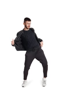 Full-length photo of a modern performer in glasses, black leather jacket, t-shirt, sports pants and light sneakers fooling around in studio. Indoor photo of an athletic person dancing and looking down isolated on white background. Music and imagination.