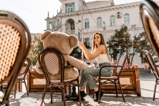 A woman sits cafe with a teddy bear next to her. The scene is set in a city with several chairs and tables around her. The woman is enjoying her time at the outdoor cafe