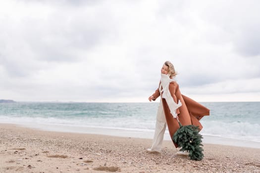 Blond woman Christmas sea. Christmas portrait of a happy woman walking along the beach and holding a Christmas tree in her hands. She is wearing a brown coat and a white suit