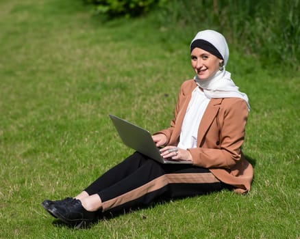 A young woman wearing a hijab sits on a lawn and uses a laptop outdoors
