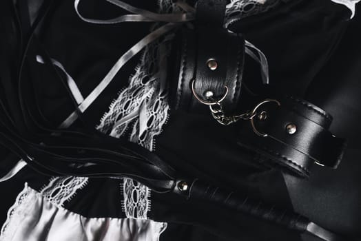 Leather handcuffs and whip for bdsm sex on black background. Maid costume for role-playing and submission