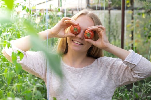 Smiling agriculture worker holding tomatoes over eyes in greenhouse. Fresh seasonal vegetables harvest. Food produce and cultivation. Sustainable farming