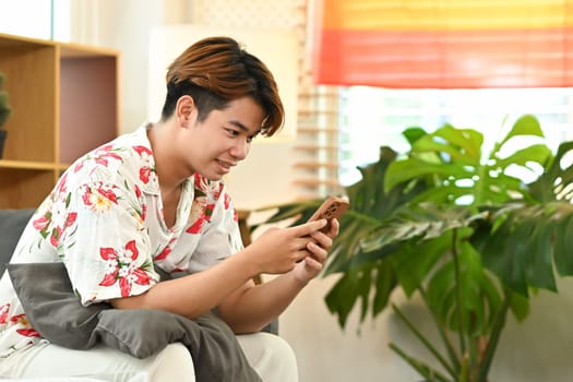 Happy young gay male reading social media post on smartphone, sitting on couch at home.