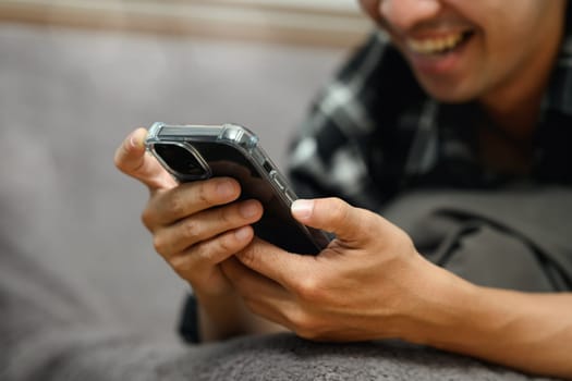 Cheerful young adult man using mobile phone while lying down on sofa at home.