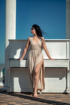 A woman in a long dress stands in front of a piano. The piano is white and has a few keys visible. The woman is posing for a photo, and the overall mood of the image is calm and serene