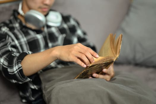 Close up of young hipster man reading book relaxing on couch at home.