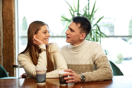 A couple sits close together at a wooden table in a brightly lit cafe, exchanging smiles over steaming cups of perhaps coffee or tea. They appear relaxed and engaged in a cheerful conversation, with natural light filtering through the space creating a warm and inviting atmosphere.