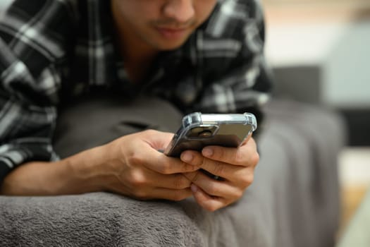 Cropped shot of young man reading messages on mobile phone lying down on sofa at home.