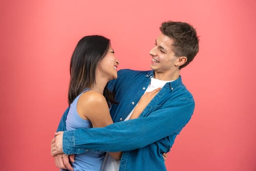 Loving young man hugging his pretty girlfriend on pink studio background, happy multiracial lovers enjoying time together, hugs, copy space.