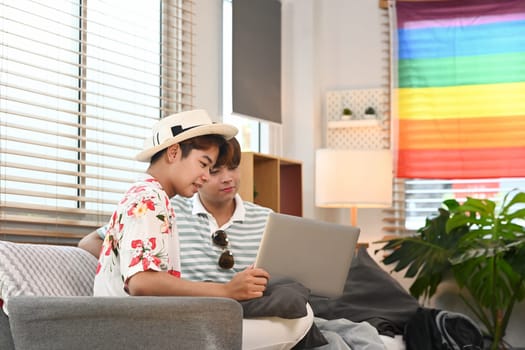 Cheerful young gay couple sitting on couch and using laptop, making hotel reservation in internet.