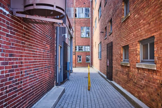 Urban alleyway in Fort Wayne with red brick buildings, industrial fire escape, and barred windows, illuminated by midday light.