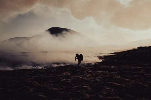 A photographer with a backpack and a camera takes a photo of flowing lava and burning moss. The ground and the background are covered with smoke of the burning moss and erupting lava. 2023 Litli-Hrutur volcano eruption, Fagradalsfjall Volcano, Reykjanes Peninsula, Iceland.