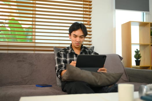Smiling Young man watching video on digital tablet while relaxing on a couch at home.