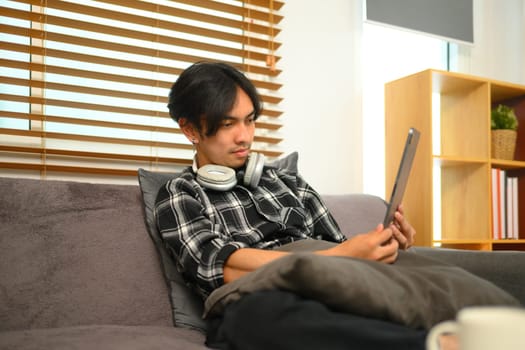 Attractive young Asian man with headphone using digital tablet in living room.