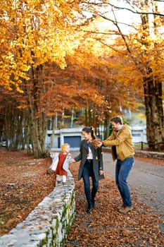 Laughing dad throws up dry leaves over mom and little girl walking through the autumn forest. High quality photo