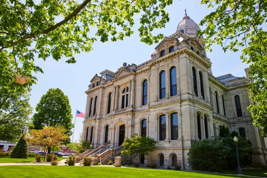 Stately Kosciusko County Courthouse in Warsaw, Indiana, framed by lush greenery and an American flag under a clear blue sky.