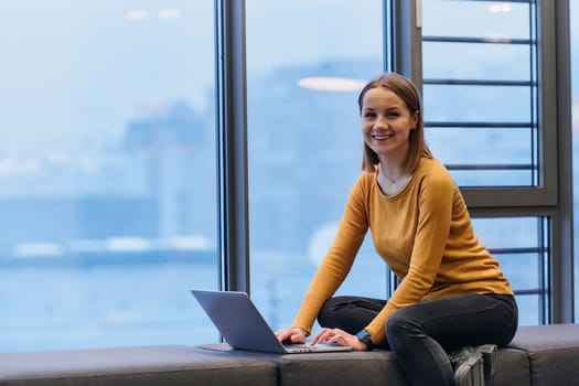 A businesswoman utilizes her laptop while seated by the window of a large corporate building, offering a picturesque view of the city skyline as her backdrop