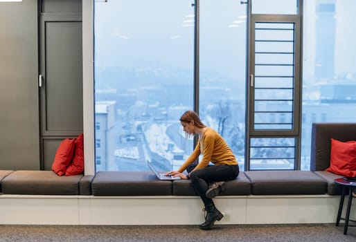 A businesswoman utilizes her laptop while seated by the window of a large corporate building, offering a picturesque view of the city skyline as her backdrop