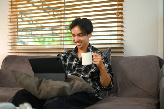 Cheerful asian man drinking coffee and using digital tablet for remote work at home.