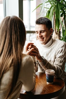 A cheerful man in a knit sweater is enjoying a chat with a woman at a wooden table in a quaint cafe. A drink in a disposable cup suggests a relaxed, informal setting, and natural light spilling from the window adds warmth to the scene. Their engagement in conversation captures a moment of genuine connection and companionship.