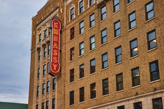 Vintage neon EMBASSY sign on a historical brick building in downtown Fort Wayne, under a cloudy sky.