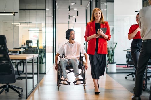 A business leader with her colleague, an African-American businessman who is a disabled person, pass by their colleagues who work in modern offices.