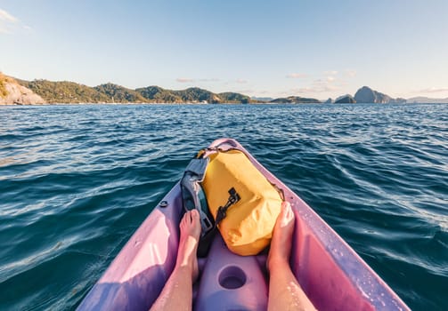 A person is kayaking in clear blue coastal waters surrounded by scenic hills and distant rocks on a sunny afternoon. Feet are visible, along with a waterproof bag on the kayak.