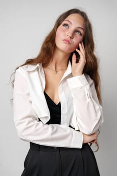 A young woman with flowing, wavy brown hair showcases her casual fashion sense, wearing an unbuttoned white shirt over a black crop top, paired with a black bottom. Her pose exudes confidence as she gazes off-camera, standing elegantly against a plain gray background.