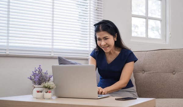 Happy Asian retired woman in her 60's enjoying social media on laptop while relaxing in living room..