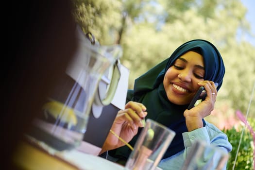 A serene African American Muslim woman in hijab engages in a peaceful conversation on her cellphone amidst the tranquil beauty of nature