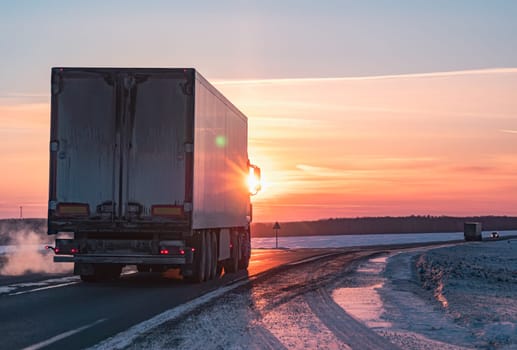 A semi truck cruises down a wintry highway as the sun sets on the horizon, casting a warm glow over the icy road and surrounding landscape.