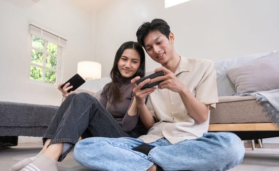 A young Asian couple happily sits together in the living room playing mobile games. Couple making romantic love in the living room.