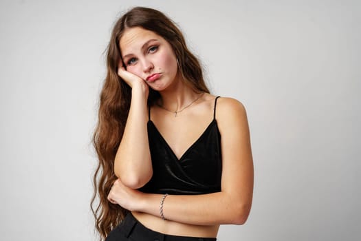 A young woman is portrayed with a bored expression, resting her cheek on hand with her elbow leaning on an invisible surface. Her long, wavy hair cascades over her shoulders and she wears a simple black top. The neutral grey backdrop emphasizes her emotive stance.