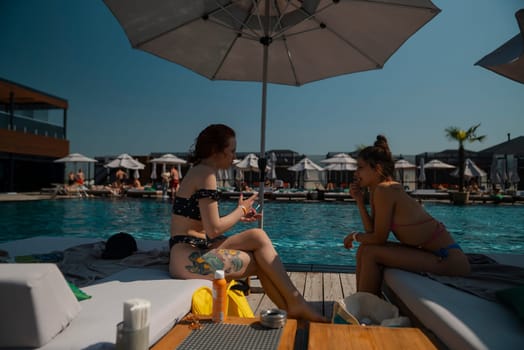 Two young women in bathing suits sit together by the pool, chatting and laughing. High quality photo