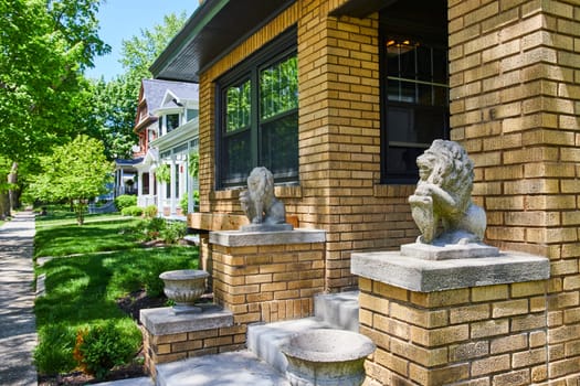 Sunny day on a peaceful suburban street in Fort Wayne, showcasing a historic yellow brick home with ornate lion statues.