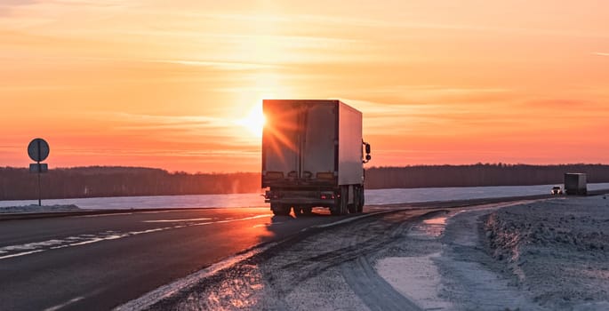 A semi truck cruises down a wintry highway as the sun sets on the horizon, casting a warm glow over the icy road and surrounding landscape.