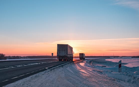 A semi truck cruises down a wintry highway as the sun sets on the horizon, casting a warm glow over the icy road and surrounding landscape.