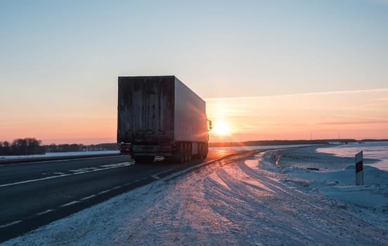A semi truck cruises down a wintry highway as the sun sets on the horizon, casting a warm glow over the icy road and surrounding landscape.
