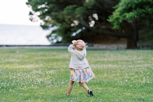 Little girl walks on a green lawn holding a cap on her head. Back view. High quality photo